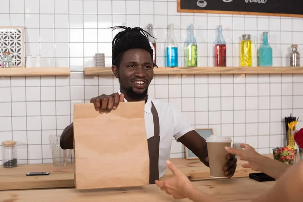 Sorrindo Africano americano barista dando xícara descartável de café e pacote kraft para o cliente — Fotografia de Stock