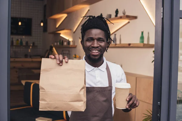 Sonriente africano americano barista celebración de taza desechable de café y kraft paquete - foto de stock