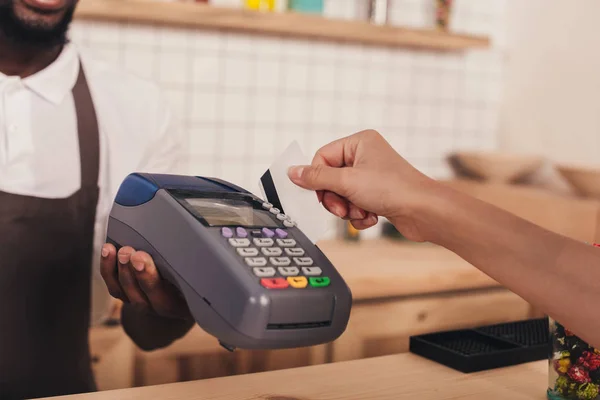 Vista recortada del cliente que paga con tarjeta de crédito y terminal en la cafetería - foto de stock