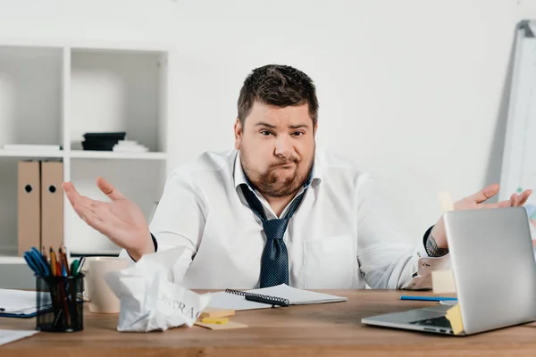 Verwirrter übergewichtiger Geschäftsmann sitzt am Arbeitsplatz mit Dokumenten und Laptop — Stockfoto