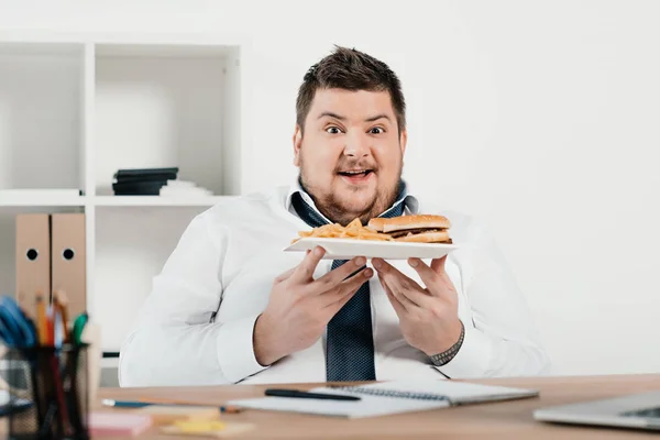 Excitado hombre de negocios con sobrepeso con hamburguesa y papas fritas en la oficina - foto de stock