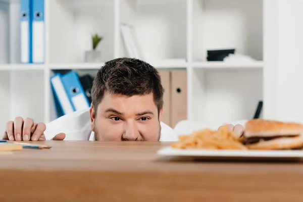 Uomo d'affari in sovrappeso guardando hamburger e patatine fritte sul posto di lavoro — Foto stock