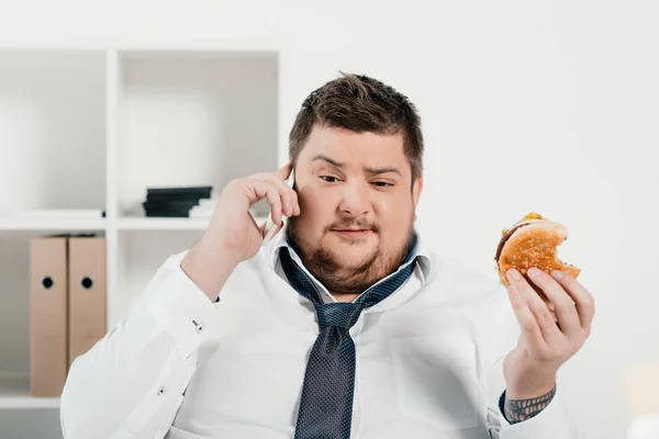 Overweight businessman talking on smartphone while eating hamburger in office — Stock Photo