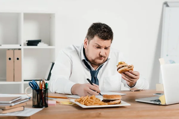 Empresário com excesso de peso trabalhando enquanto come hambúrguer e batatas fritas no escritório — Fotografia de Stock