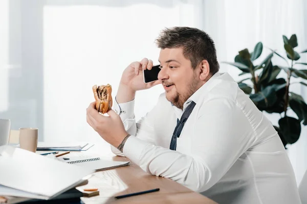 Homme d'affaires en surpoids parlant sur smartphone tout en mangeant un hamburger au bureau — Photo de stock