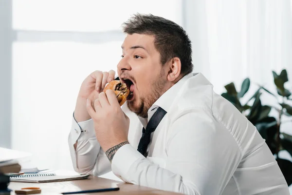 Hombre de negocios gordo hablando en el teléfono inteligente mientras come hamburguesa en la oficina - foto de stock