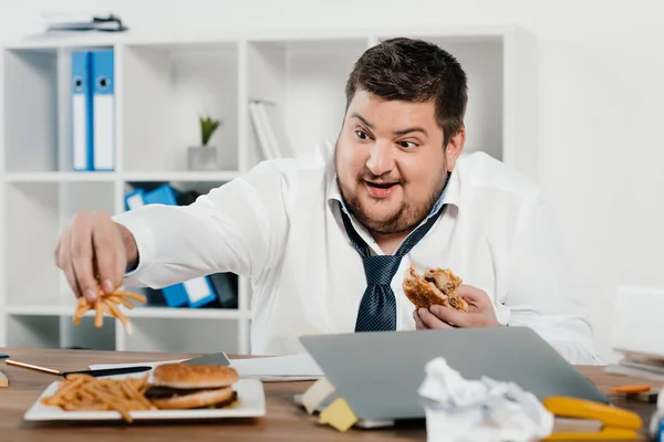 Overweight businessman eating hamburgers and french fries in office — Stock Photo