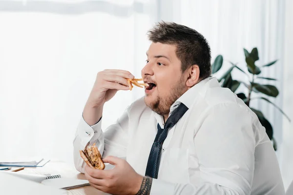 Overweight businessman eating hamburger and french fries at workplace — Stock Photo