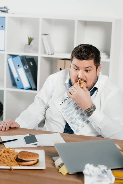 Gordo hombre de negocios comiendo hamburguesas y papas fritas en la oficina - foto de stock