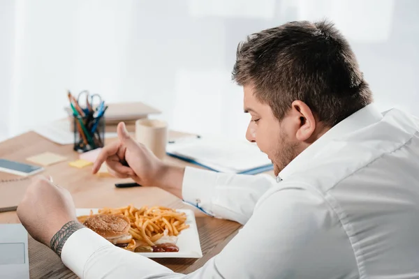Overweight businessman having lunch with hamburger and french fries in office — Stock Photo