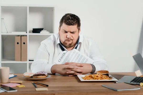Confundido hombre de negocios con sobrepeso eligiendo donas o hamburguesa con papas fritas - foto de stock
