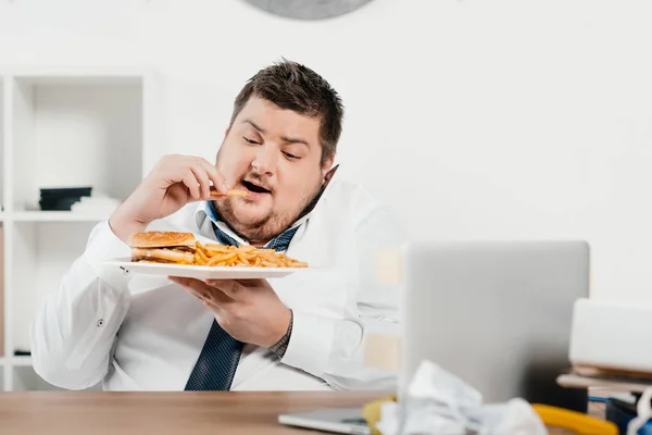 Overweight businessman having lunch with hamburger and french fries in office — Stock Photo