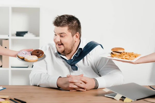 Hombre de negocios con sobrepeso eligiendo rosquillas o hamburguesa con papas fritas para el almuerzo - foto de stock