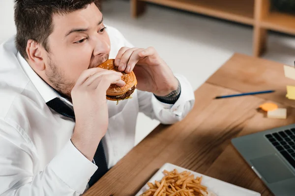 Overweight businessman eating hamburger at workplace — Stock Photo