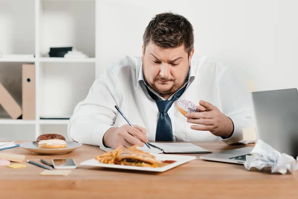 Empresário com excesso de peso comer donuts, hambúrguer e batatas fritas enquanto acorda no escritório — Fotografia de Stock