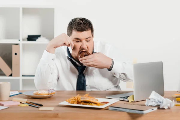 Hombre de negocios con sobrepeso comiendo rosquillas, hamburguesas y papas fritas en el lugar de trabajo - foto de stock