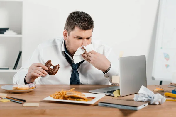 Hombre de negocios con sobrepeso comiendo donas, hamburguesa y papas fritas mientras se despierta con el ordenador portátil en la oficina - foto de stock