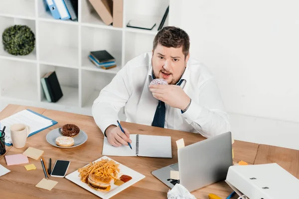 Uomo d'affari sovrappeso mangiare ciambelle, hamburger e patatine fritte mentre si sveglia in ufficio — Foto stock