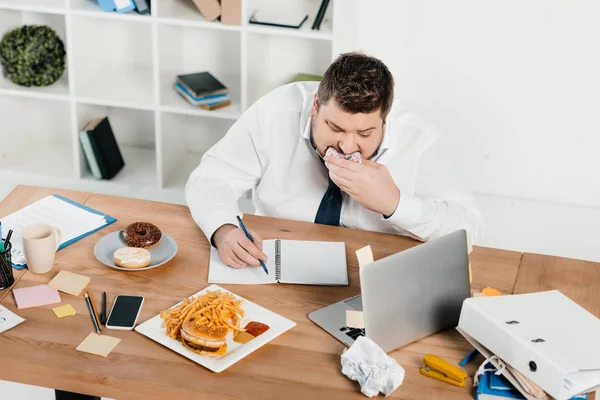 Empresário com excesso de peso comer donuts, hambúrguer e batatas fritas enquanto acorda no escritório — Fotografia de Stock