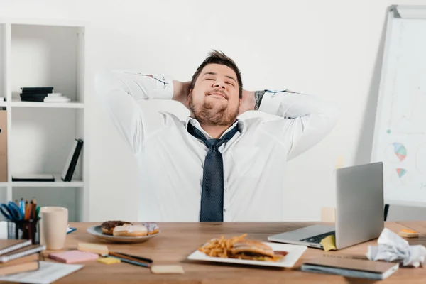 Hombre de negocios con sobrepeso relajándose en el lugar de trabajo con computadora portátil y comida chatarra - foto de stock