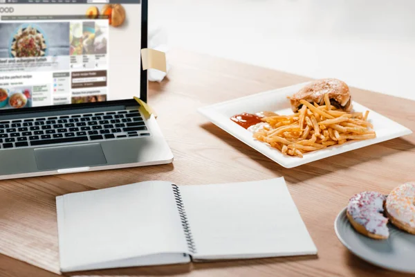 Portátil con sitio web, bloc de notas, donas y hamburguesa con papas fritas en el lugar de trabajo - foto de stock