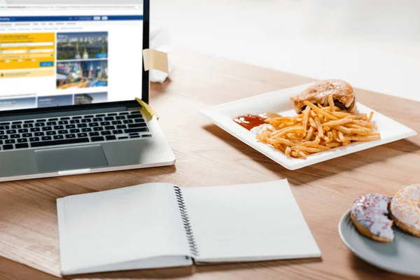 Portátil con sitio web de reserva, bloc de notas, donas y hamburguesa con papas fritas en el lugar de trabajo - foto de stock