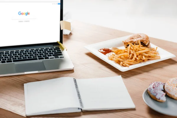 Portátil con google sitio web, bloc de notas, donas y hamburguesa con papas fritas en el lugar de trabajo - foto de stock