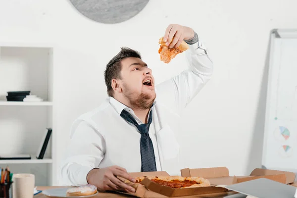 Hombre de negocios con sobrepeso comiendo pizza para el almuerzo en el lugar de trabajo - foto de stock