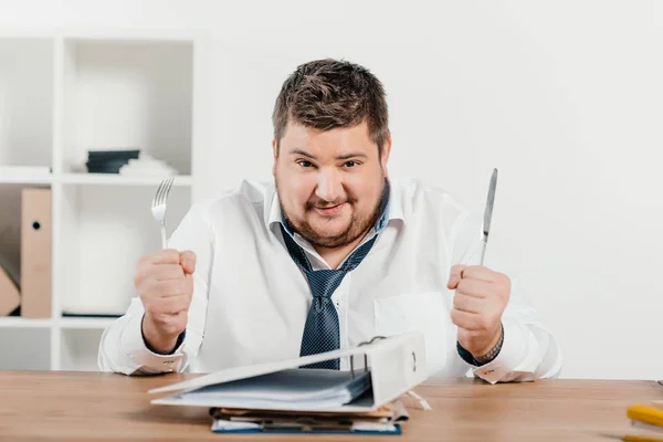 Übergewichtiger Geschäftsmann mit Gabel und Messer sitzt mit Ordnern am Tisch — Stockfoto