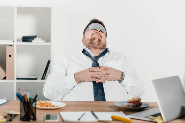 Homme d'affaires en surpoids en masque de sommeil relaxant sur le lieu de travail — Photo de stock