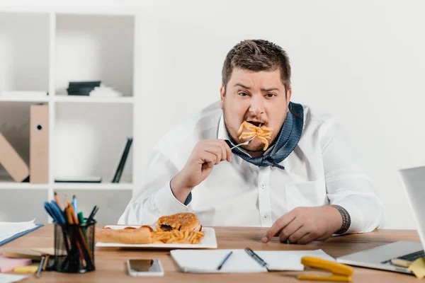 Empresario con sobrepeso comiendo comida chatarra en el lugar de trabajo - foto de stock