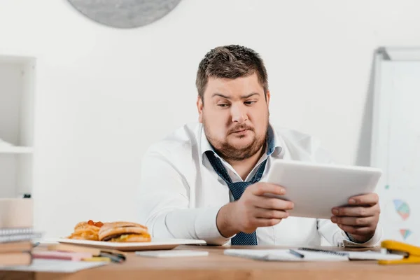 Hombre de negocios con sobrepeso mirando comida chatarra mientras trabaja con la tableta en el lugar de trabajo - foto de stock