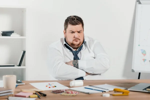 Overweight businessman sitting at workplace and looking at pills — Stock Photo