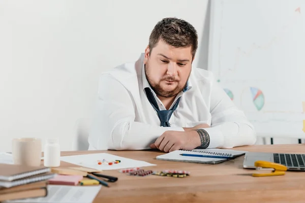 Hombre de negocios con sobrepeso sentado en el lugar de trabajo y mirando pastillas — Stock Photo