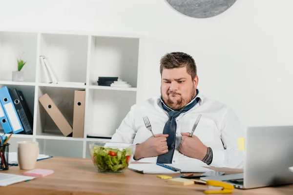 Homme d'affaires en surpoids confus avec fourchette et couteau regardant la salade fraîche — Photo de stock