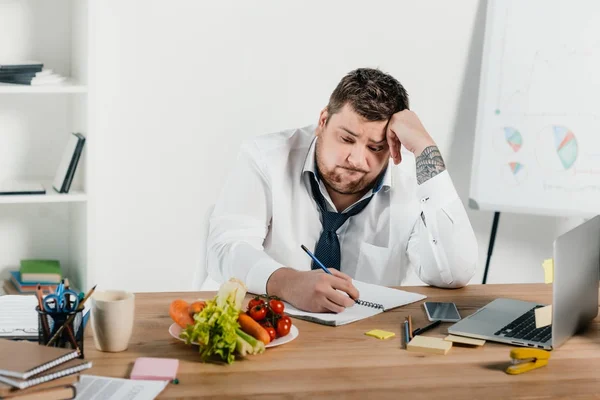 Upset overweight businessman looking at vegetables while working in office — Stock Photo