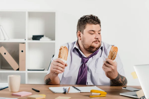 Hombre de negocios con sobrepeso comiendo perritos calientes en el lugar de trabajo en la oficina - foto de stock