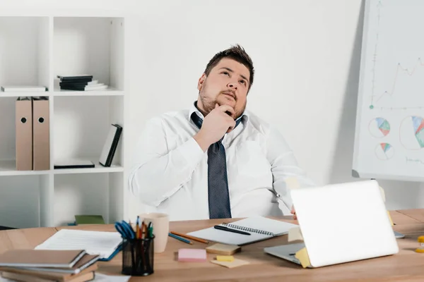 Nachdenklicher übergewichtiger Geschäftsmann arbeitet im Büro mit Notizblock und Laptop — Stockfoto