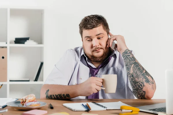 Hombre de negocios tatuado con sobrepeso hablando en el teléfono inteligente en el lugar de trabajo - foto de stock