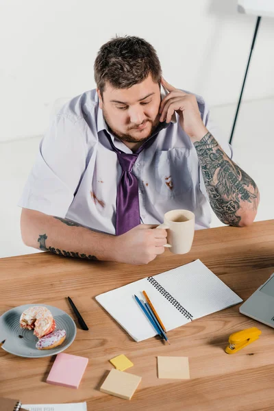 Hombre de negocios tatuado con sobrepeso hablando en el teléfono inteligente en el lugar de trabajo - foto de stock