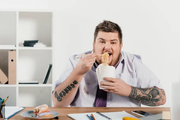 Übergewichtiger tätowierter Geschäftsmann isst Nudeln am Arbeitsplatz im Büro — Stockfoto