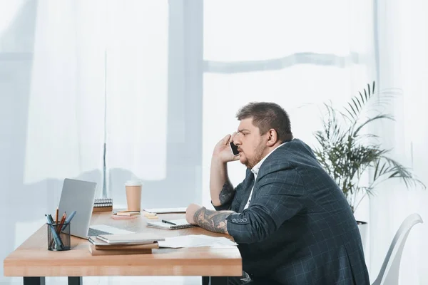 Hombre de negocios con sobrepeso hablando en el teléfono inteligente mientras trabaja con el ordenador portátil en la oficina - foto de stock