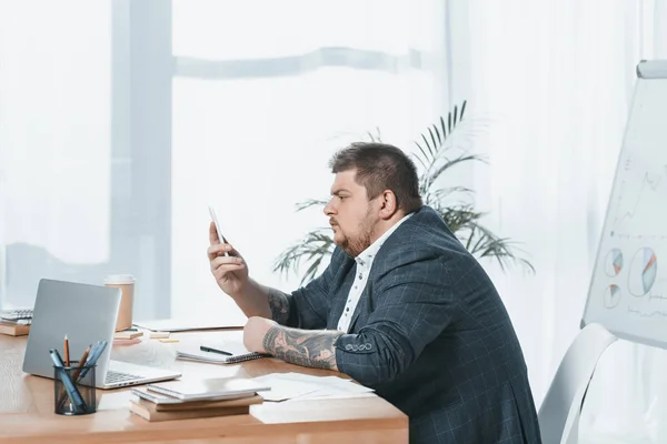 Homem de negócios com sobrepeso em terno trabalhando com smartphone e laptop no local de trabalho — Fotografia de Stock