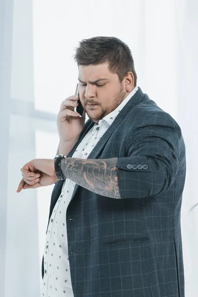 Overweight businessman looking at watch while talking on smartphone — Stock Photo