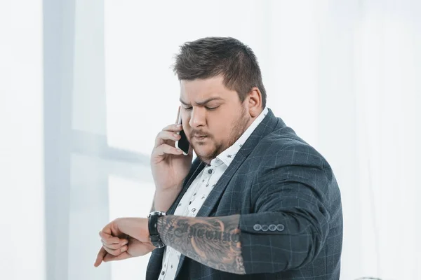 Hombre de negocios con sobrepeso mirando el reloj mientras habla en el teléfono inteligente en la ventana - foto de stock