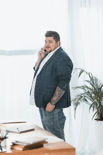 Hombre de negocios con sobrepeso hablando en el teléfono inteligente en la ventana en la oficina - foto de stock