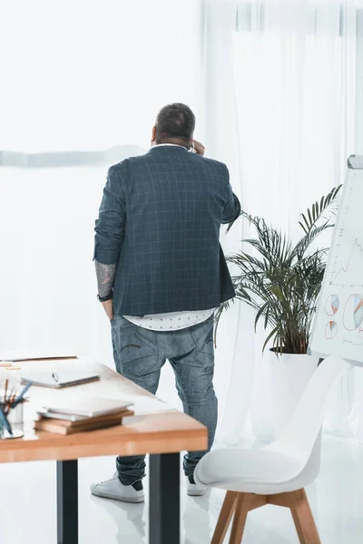 Rückansicht eines übergewichtigen Geschäftsmannes, der am Fenster im Büro mit dem Smartphone spricht — Stockfoto