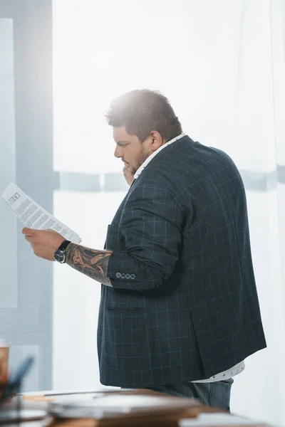 Übergewichtiger Geschäftsmann im Anzug schaut sich Dokument an, während er im Büro am Fenster steht — Stockfoto