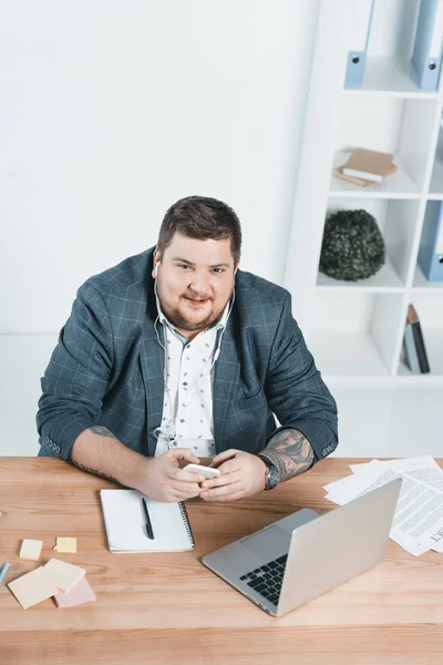 Hombre de negocios con sobrepeso escuchando música y trabajando con computadora portátil en el lugar de trabajo - foto de stock