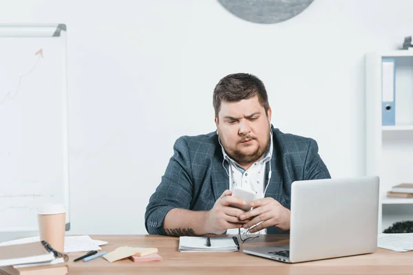 Homem de negócios com sobrepeso ouvindo música com smartphone enquanto usa laptop no local de trabalho — Fotografia de Stock